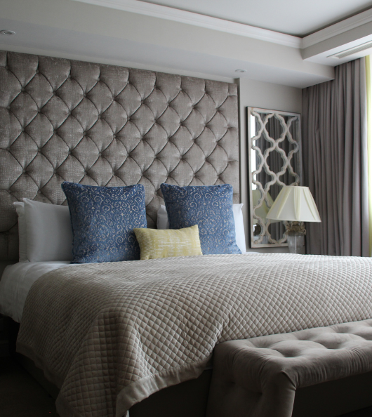 A master bed with grey headboard, grey sheets, a grey ottoman, and blue & yellow cushions